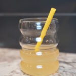 Bees Knees yellow cocktail in a bubble "bee hive resembling" cup. Yellow heart shaped straw inside. Cup is sitting on a granite counter in front of a grey backdrop.