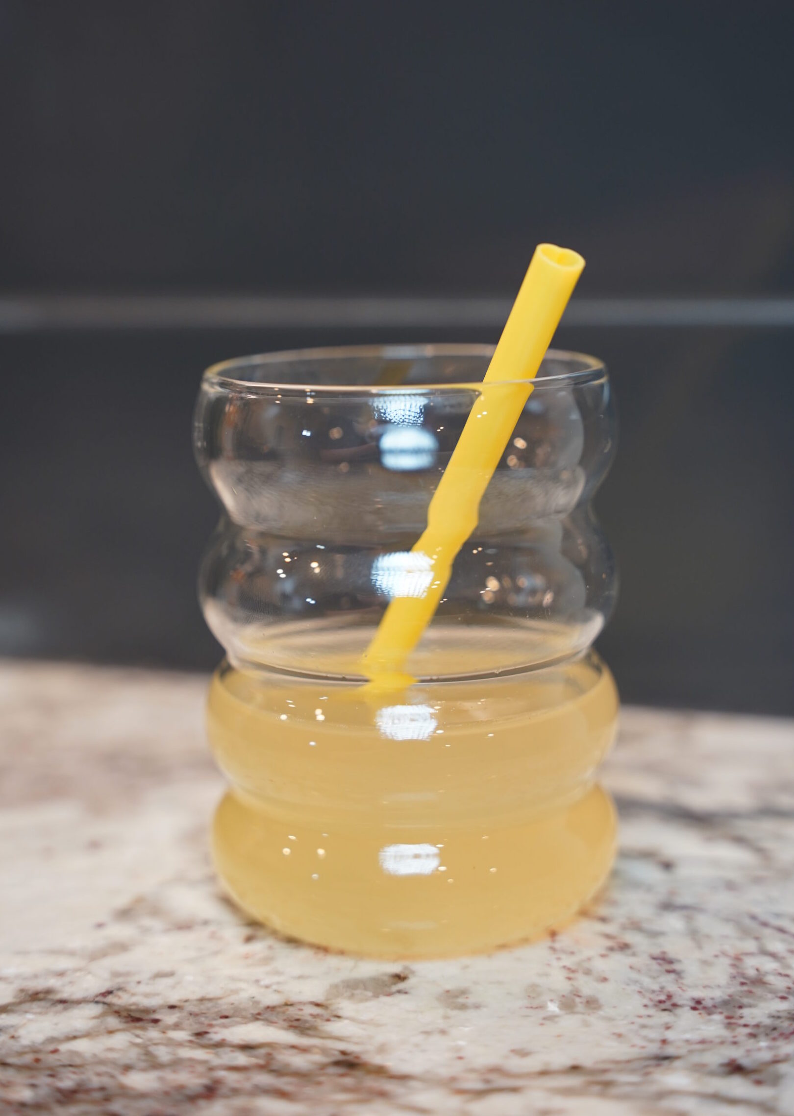 Bees Knees yellow cocktail in a bubble "bee hive resembling" cup. Yellow heart shaped straw inside. Cup is sitting on a granite counter in front of a grey backdrop.