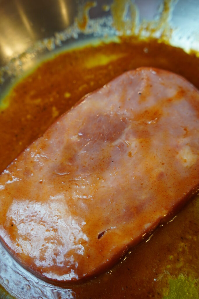 Ham steak sitting in a stainless steel pan with glaze ingredients, before glaze thickens and caramelizes to meat.