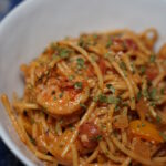 Cajun shrimp pasta in a white bowl topped with finely minced parsley. deep red sauced noodles with boiled shrimp, onion, bell pepper, garlic and seasonings.