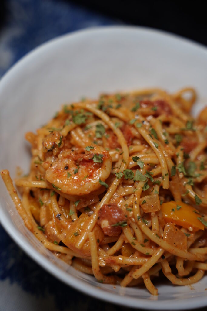 Cajun shrimp pasta in a white bowl topped with finely minced parsley. deep red sauced noodles with boiled shrimp, onion, bell pepper, garlic and seasonings.