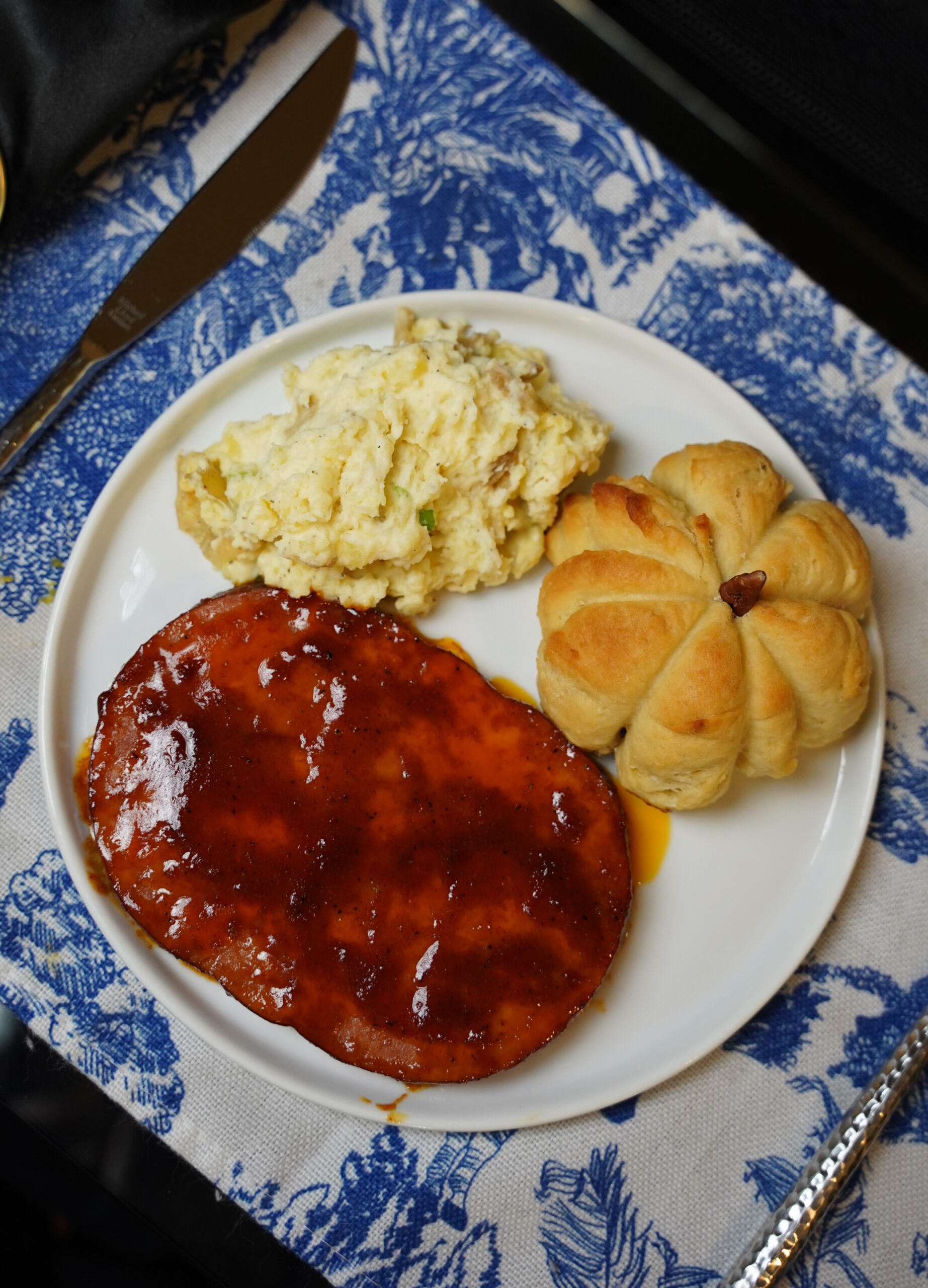 Brown Sugar Dijon Glazed Ham Steak, Pumpkin Shaped Dinner Roll, and Chef Level Mashed Potatoes on a white plate and blue and white place mat.