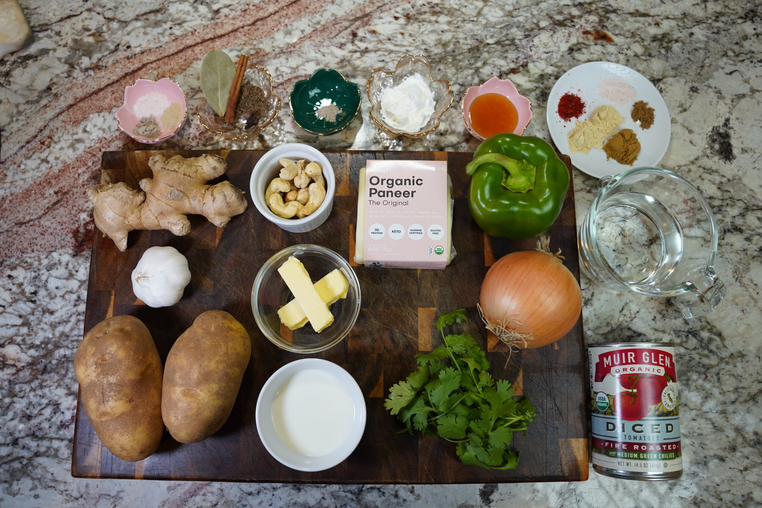 Malai Kofta ingredients on a wooden cutting board and granite countertop. potato, onion, green bell pepper, cilantro, ginger, garlic, cashews, paneer, butter, heavy cream, diced fire roasted tomato with green chili, honey, and spices