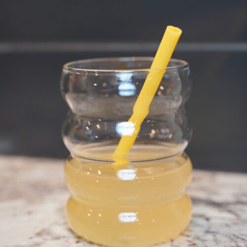 Bees Knees yellow cocktail in a bubble "bee hive resembling" cup. Yellow heart shaped straw inside. Cup is sitting on a granite counter in front of a grey backdrop.
