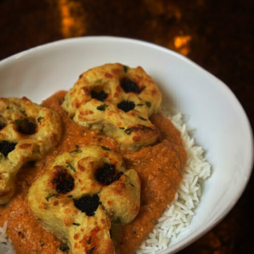 Skull Shaped Malai Kofta dish by Dakota-Delights. Jasmine rice topped with tomato curry and golden potato paneer shaped into a skull on the top. In a white bowl on a copper table.