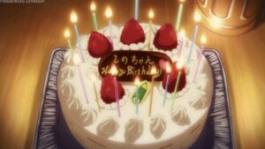 A white birthday cake with plenty of candles and strawberries sitting on top of cream. Brown cookie with "happy birthday" etched in the center.